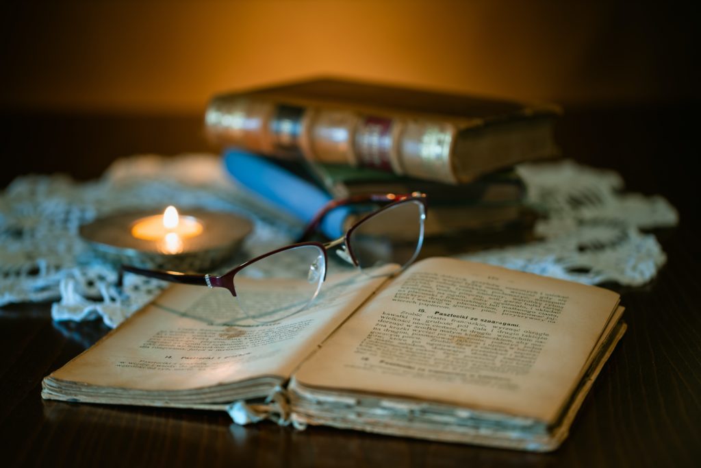 Table wiith old books and tealight on cloth. Spectacles lying on open book in front.